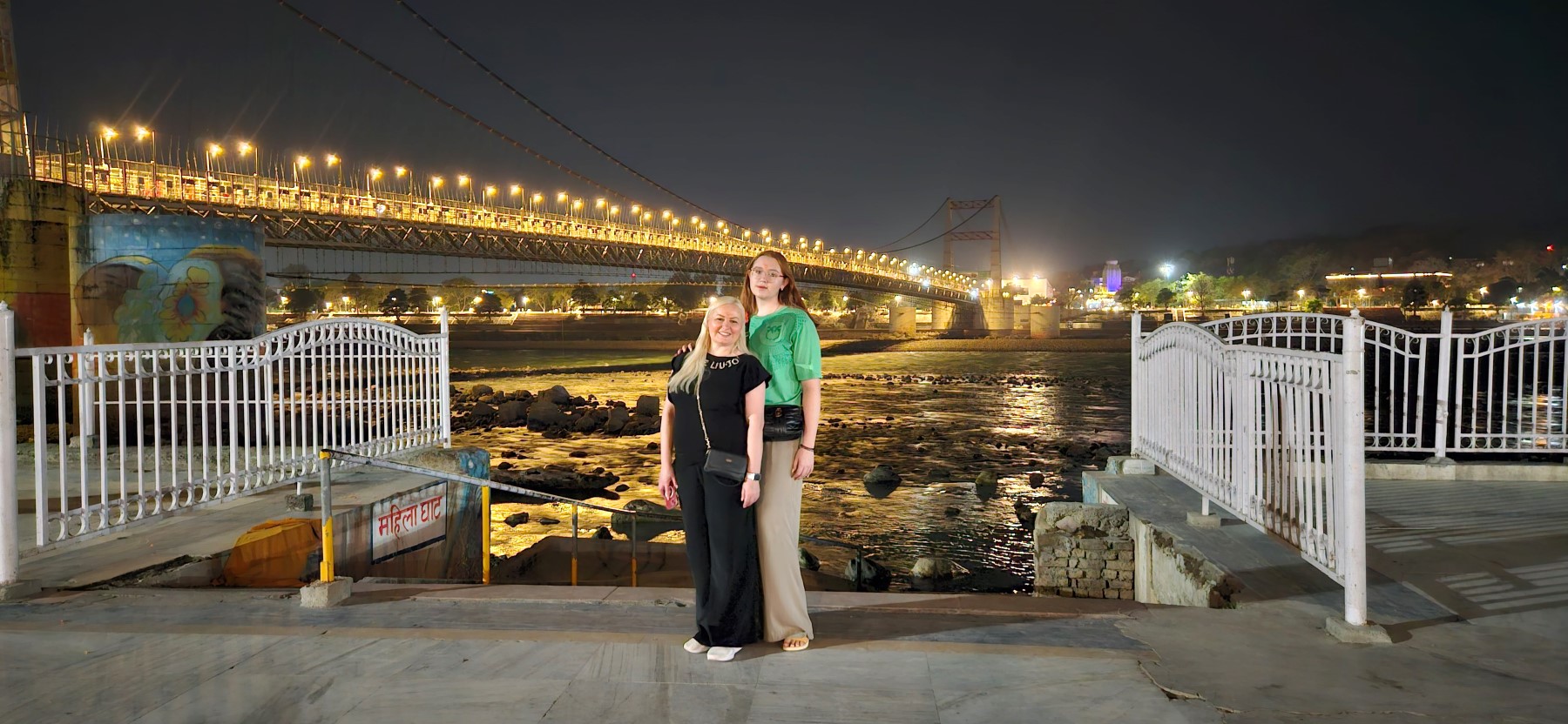 Two guests by the Ganga at night near a lit suspension bridge on the Rishikesh ghats