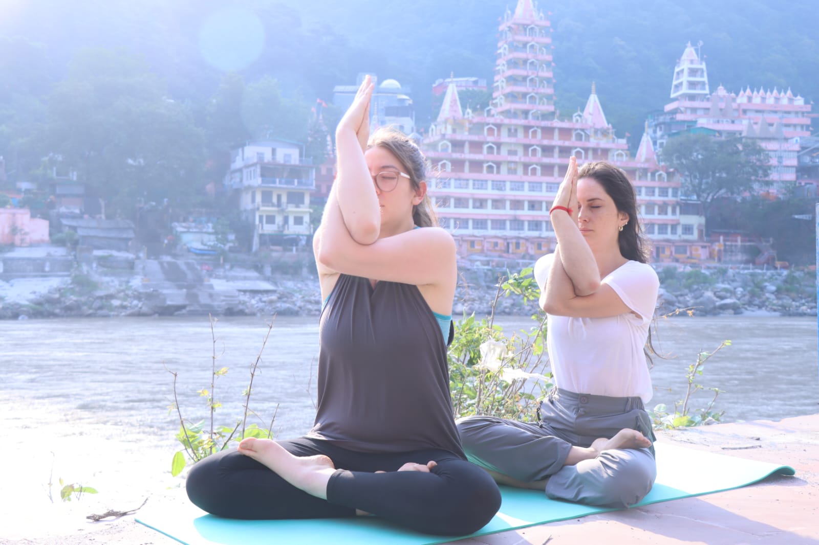 Two guests practicing yoga by the Ganga with a riverside temple behind them