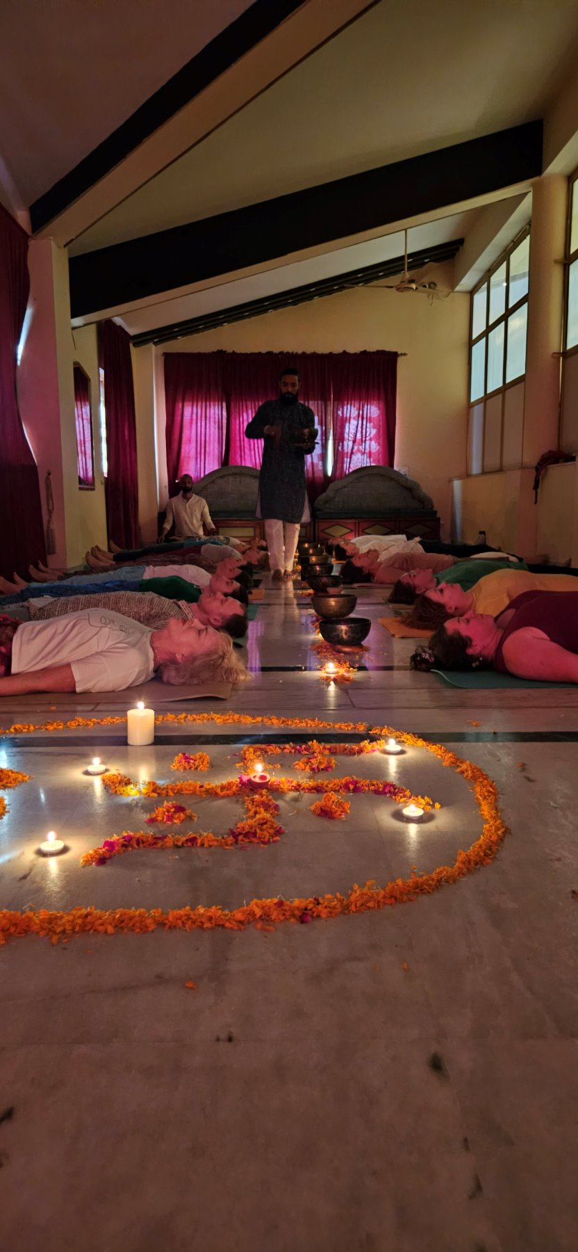 A sound healer moving between singing bowls as the group lies in savasana around a marigold Om rangoli