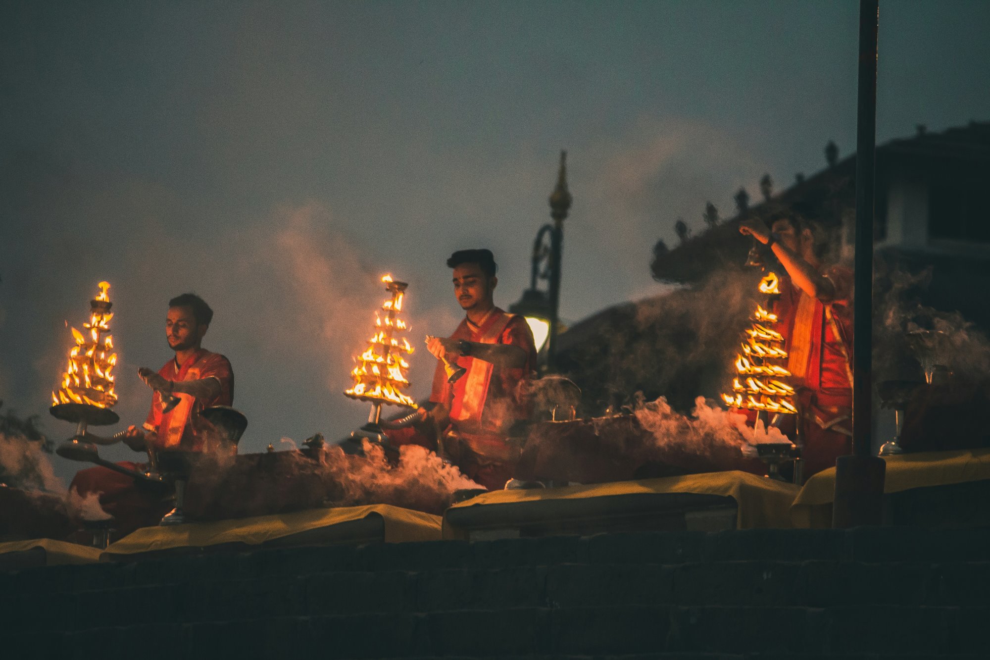 Priests performing the Ganga aarti at dusk, tiered flame lamps lit and smoke rising over the river